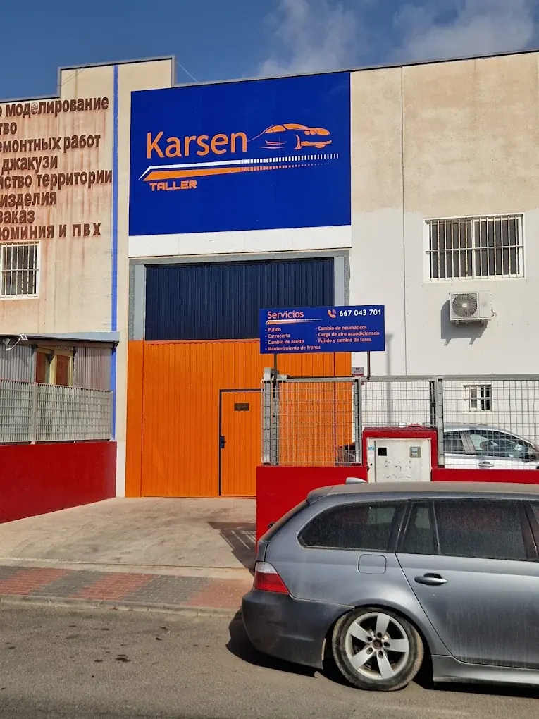 Entrada de Karsen Taller: cartel azul con el logotipo, puerta naranja y cartel de servicios con el teléfono 667 043 701