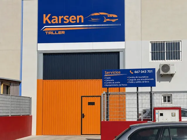 Entrada de Karsen Taller: cartel azul con el logotipo, puerta naranja y cartel de servicios con el teléfono 667 043 701